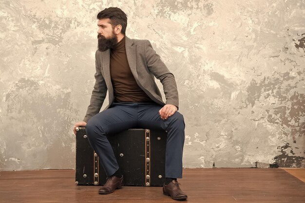 Bearded man sitting on vintage suitcase in brown blazer and turtleneck, wearing dark pants and leather shoes against a textured wall.