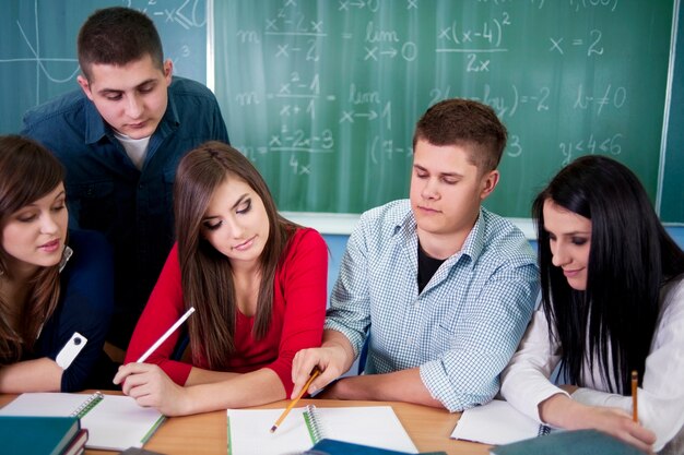 Group of students studying together at a desk, writing in notebooks with a chalkboard of math equations behind them.
