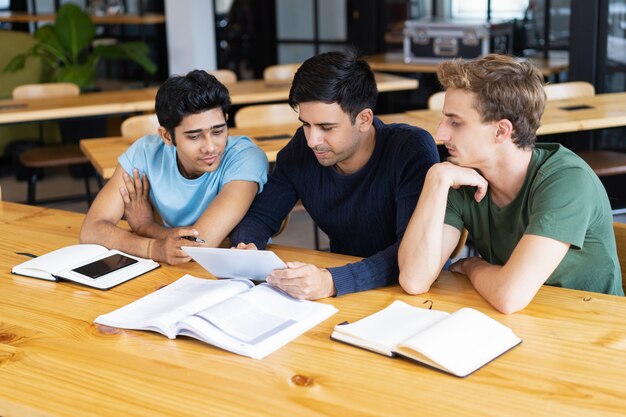 Three college students reviewing notes together at a wooden table with open books and notebooks.