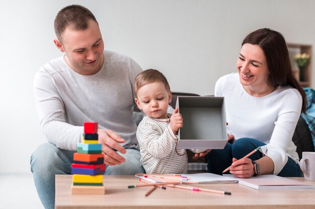 Parents playing with their toddler at a table with colorful stacking blocks and drawing supplies.