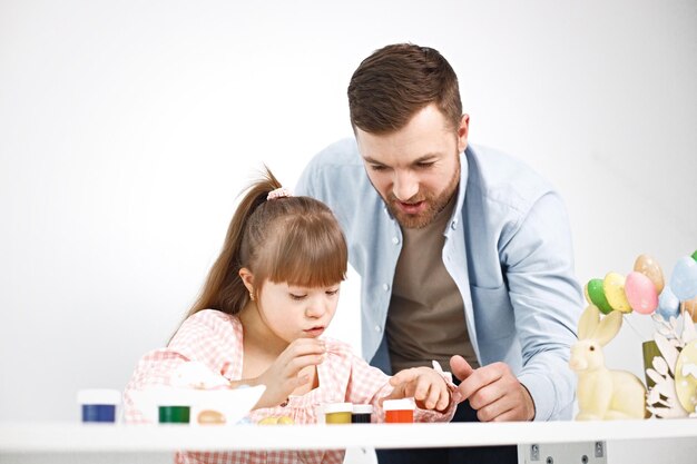 Father helping his young daughter paint at a table with colorful paints and craft supplies.