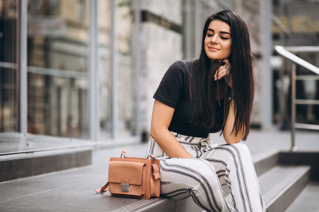 Woman in white blouse with red earrings leaning on a column, holding a patterned handbag outdoors.